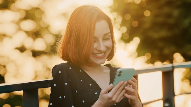Привлекательная рыжая девушка подросток женщина стоит на улице на открытом воздухе солнечные лучи закат смотрит смартфон