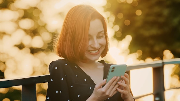 Attractive redhead girl teenager woman stands street outdoors sunlight rays sunset looks smartphone