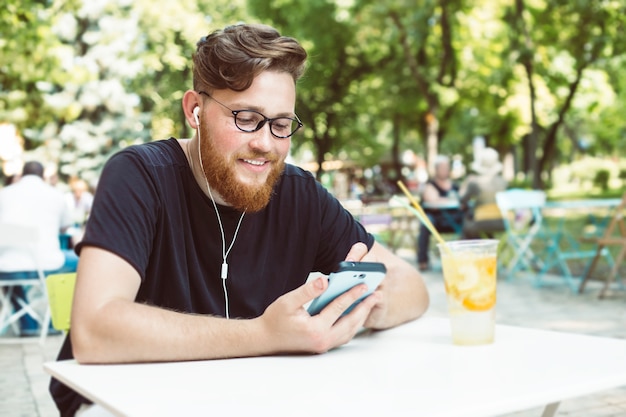 Attractive redhead man with a beard listens to music on a mobile phone while sitting at a cafe table.