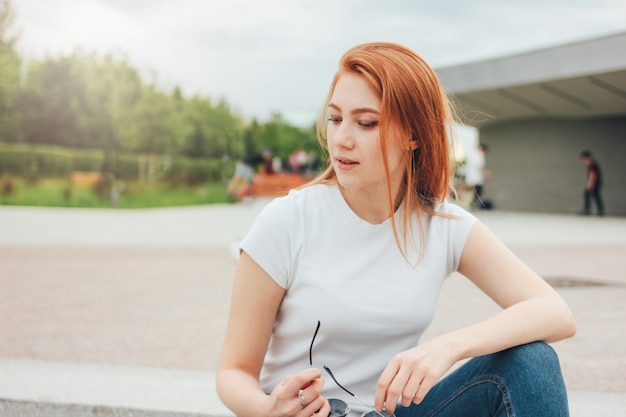 Attractive redhead smiling girl in round sunglasses in casual clothes sitting on street in city