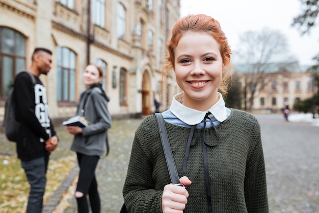 キャンパスの外に立っているバックパックを持つ魅力的な赤毛の女性の学生