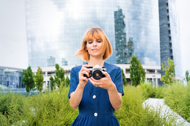 Attractive redhead woman taking photos on her camera in town against a backdrop of greenery and a modern glass tower block on a breezy summer day