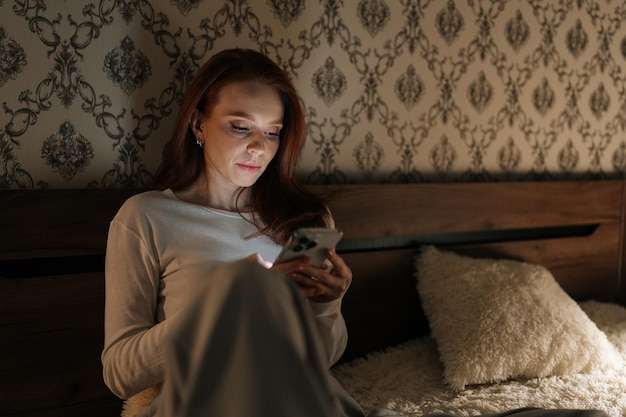 attractive redhead young woman relaxing using smartphone sitting in bed at night looking to screen