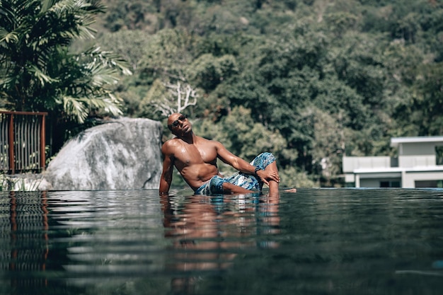 Привлекательный и расслабленный арабский мужчина без рубашки в бирюзовом саронге, лежащий у пейзажного бассейна в солнечный день; отражение в воде, пальмы и отель на заднем плане; концепция отпуска.