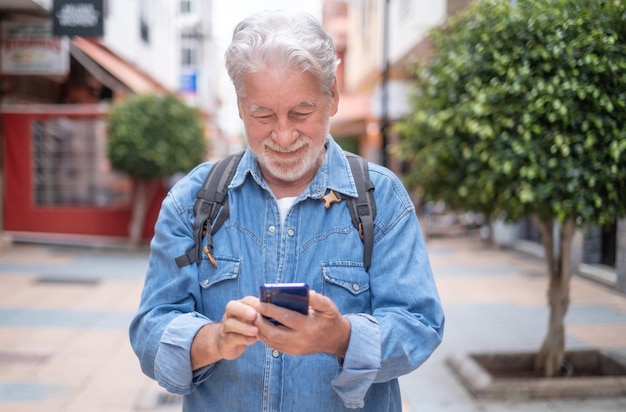 Attractive relaxed mature adult Caucasian man white- haired walking in city street holding cellphone in hand. Beautiful senior man denim dressed enjoys free time and smiles looking at smart phone