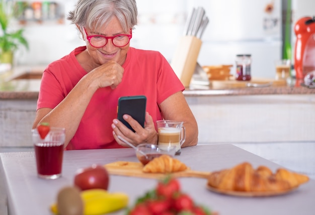 Attractive retired senior woman enjoying breakfast at home with croissant and cappuccino. Looking at news on mobile phone