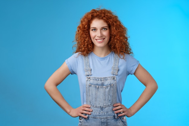 Attractive sassy motivated redhead european female curly-haired hold hands waist empowered express confidence self-assured look forward ready win conqueror, stand blue background. Copy space