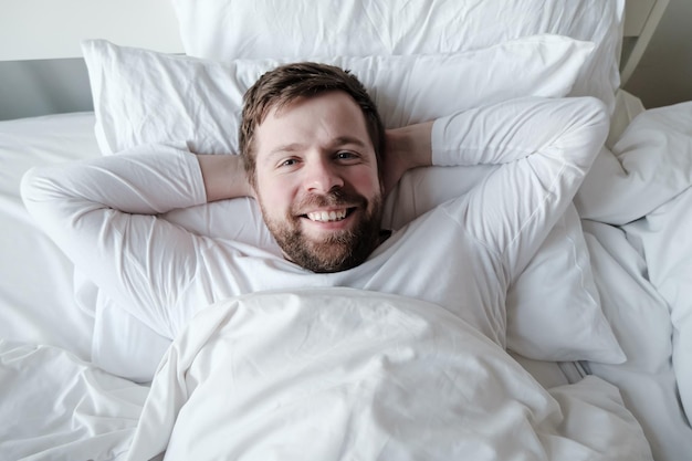 Photo attractive satisfied man in pajamas lies in bed on a soft pillow with hands under head