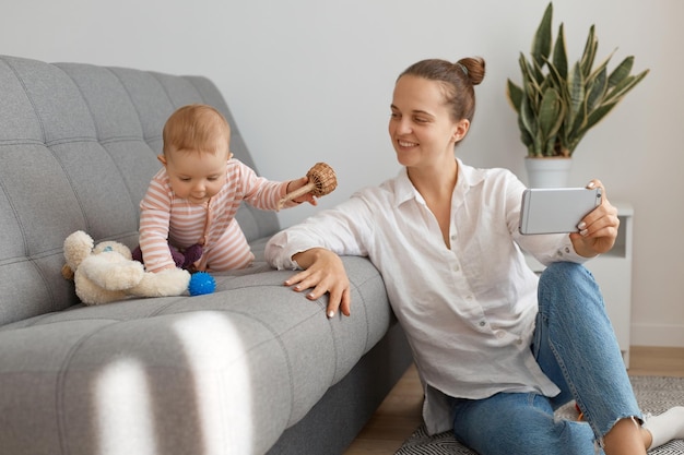 Attractive satisfied woman wearing white shirt and jeans sitting on floor and holding cell phone looking at her baby daughter crawling on cough mother smiling expressing happiness