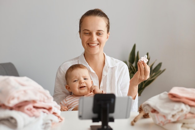 Attractive satisfied young adult woman sitting at table with toddler kid and recording video for her vlog, advertising good nipples for babies, making content with her daughter.