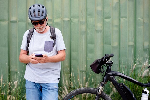 Attractive senior man with helmet and backpack using mobile\
phone while enjoying sport activity with electric bicycle in\
outdoors sustainable mobility concept