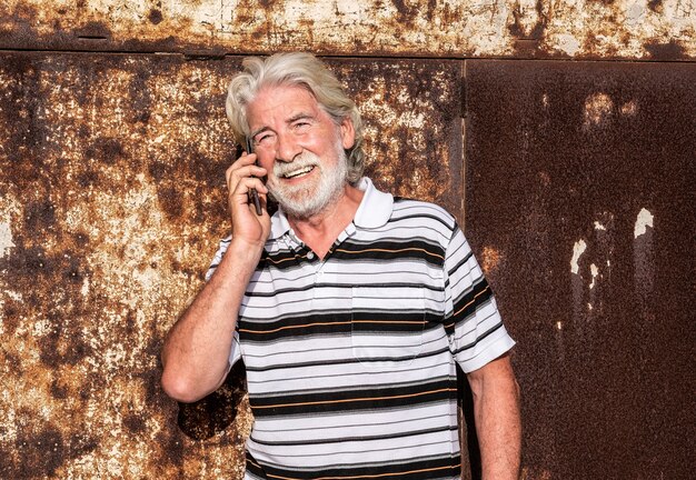 Attractive senior people male with gray hair smiling and talking by cell phone. leaning against a rusted iron panel. Sunset light.