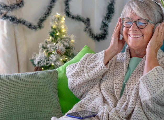 魅力的な年配の女性は、ヘッドフォンで音楽を聴きながら、自宅のソファでリラックスして幸せに笑います。背景のクリスマスの装飾