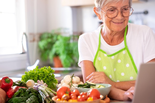 Donna anziana attraente che prepara le verdure nel menu di ricerca della cucina di casa al computer portatile anziani caucasici che godono di un'alimentazione sana