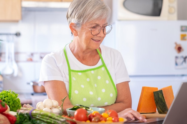 Attractive senior woman preparing vegetables in the home\
kitchen searching menu at laptop computer caucasian elderly people\
enjoying healthy eating