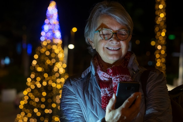 彼女のスマートフォンでメッセージを読んで笑顔で夜に立っている魅力的な年配の女性。黄色い電球のライトで飾られたクリスマスツリーの前