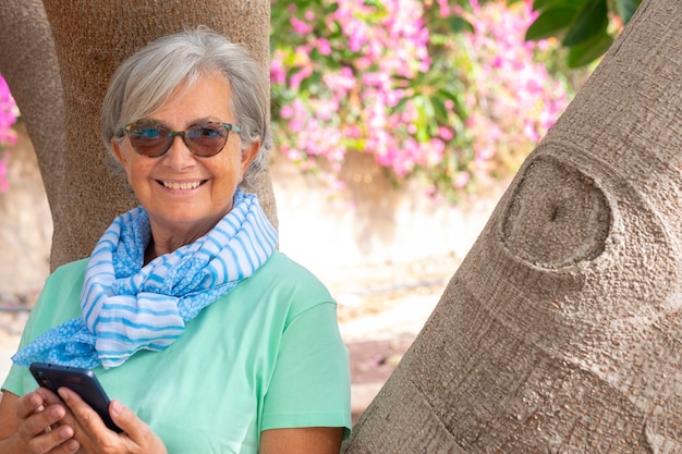 Attractive senior woman white haired looking at camera wearing sunglasses and fashion scarf using smart phone and smiling. Standing in public park against a tree trunk with beautiful pink flowers