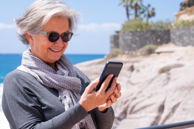 海でスマホを使って笑顔で白髪の魅力的な年配の女性。うれしそうなライフスタイルのコンセプト、幸せな引退