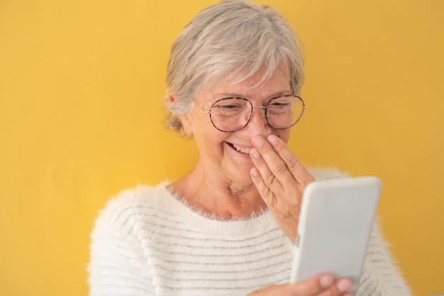 Attractive senior woman in white sweater laughing amused while\
using phone in video chat old generation and new technology