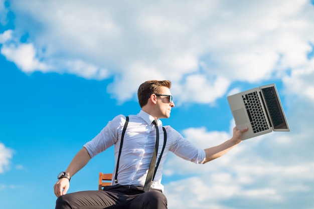 Attractive serious busunessman in white shirt, tie, braces and sunglasses going to break the laptop