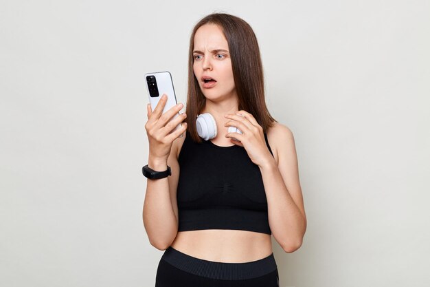 Attractive shocked surprised mslim woman with headphones wearing sportswear posing against gray background holding mobile phone looking at display with astonished face