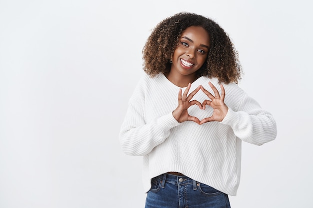 Attractive silly african-american woman afro hairstyle show heart gesture tilt head smiling joyfully express sympathy passion romantic attitude, standing white wall carefree