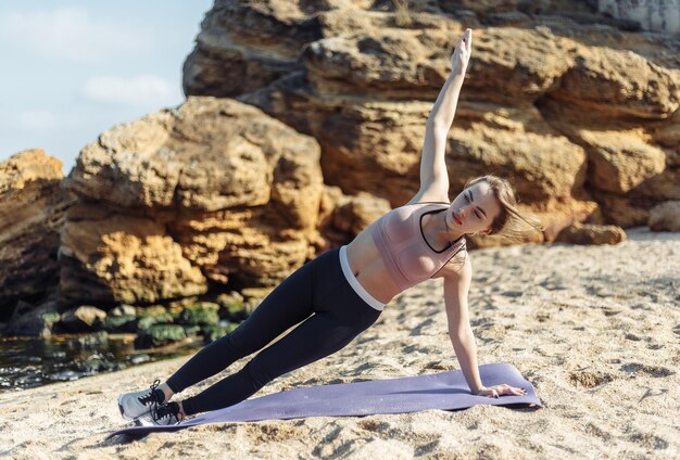 Attractive slim fit woman practicing side plank exercise on wild beach at bright sunny day