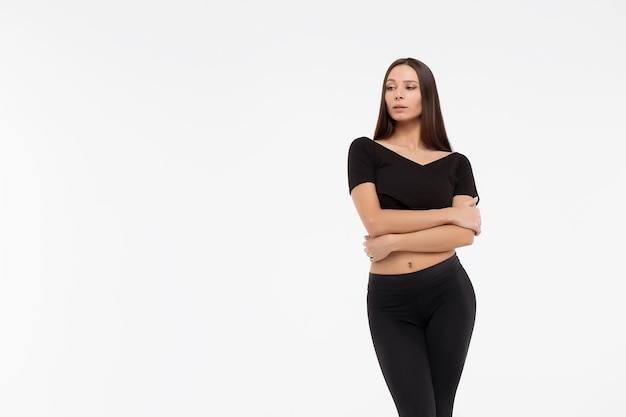 Attractive slim lady in black top and leggings embracing body and looking at camera