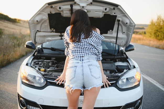Photo attractive slim young girl in summer shorts and shirt repairs a broken car a beautiful woman stands near raised car hood