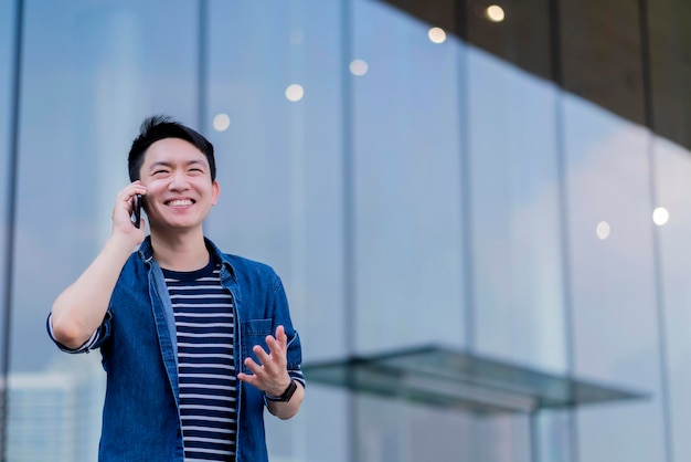 Привлекательный умный азиатский внештатный мужчина в повседневной одежде улыбается веселой уверенной беседой со смартфоном за пределами концепции строительных технологий