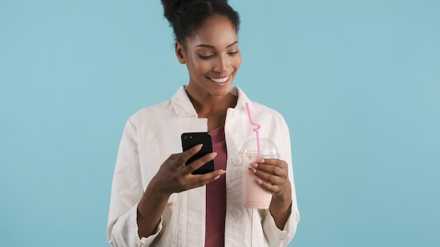 Attractive smiling african american girl happily using smartphone and drinking milkshake over colorful background