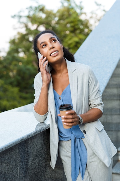 Attractive smiling african business woman standing outdoors at the city, leaning on stairs, holding takeaway coffee cup, talking on mobile phone