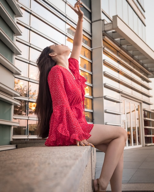 An attractive smiling Asian girl in red sits on a granite dais among glass buildings.