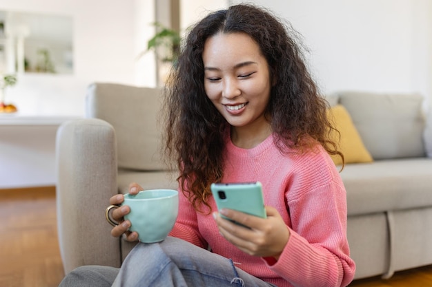 Attraente donna asiatica sorridente che usa lo smartphone mentre si siede sul divano di casa concetto di comunicazione e videochiamata