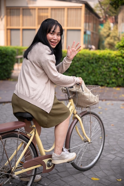 An attractive smiling Asian woman waves her hand to greet someone while riding a bike in the city