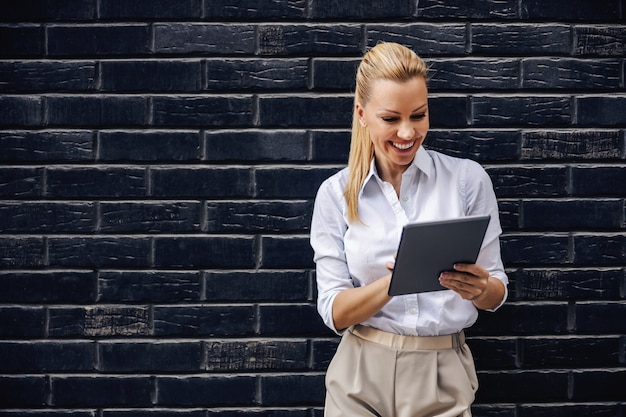 Attractive smiling blond fashionable businesswoman standing in front of the brick wall and using tablet.