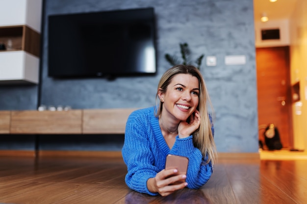 Attractive smiling blond young woman lying down on stomach in living room and using smart phone to hang on social media sites