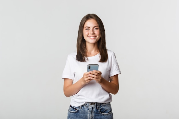 Attractive smiling brunette girl texting message, using mobile phone and looking at camera.