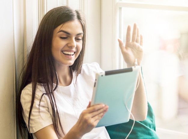 Attractive smiling brunette girl wears white tshirt using tablet computer for videochat with friends sitting on the windwsill