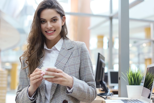 Attraente imprenditrice sorridente in piedi in ufficio con una tazza di caffè.