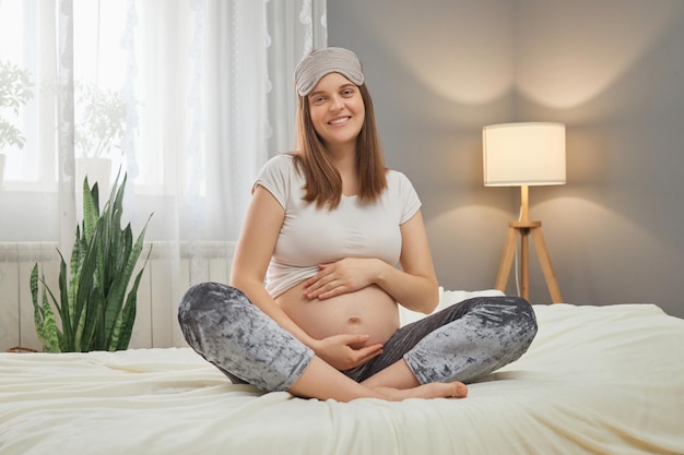 Attractive smiling cheerful Caucasian pregnant woman sitting in bed and holding her belly looking at camera with happy smile enjoying her pregnancy stroking tummy