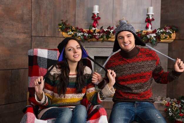 Attractive smiling couple in funny knitted Nordic hats sitting in a rocking chair near decorated fireplace in the living room