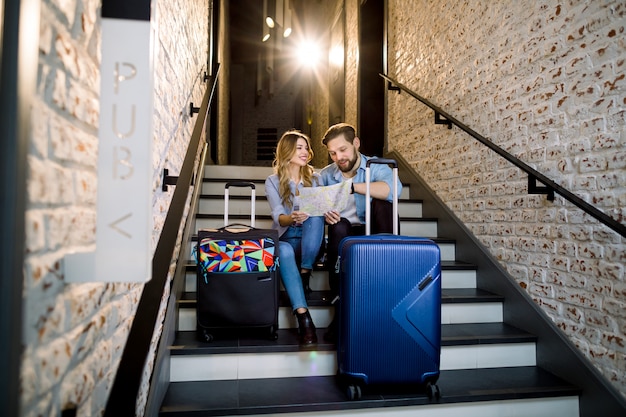 Attractive smiling couple of man and woman in smart casual business style with two suitcases, sitting on stairs in stylish loft hotel hall indoors, looking together on city map