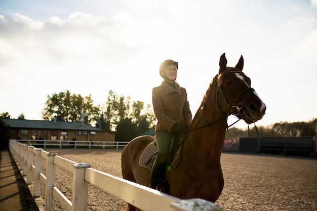 Attractive smiling female horse rider training in outdoor paddock. Riding club and equine caring concept