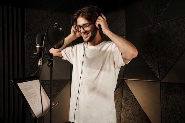 Attractive smiling guy happily wearing headphones recording new song in modern sound studio