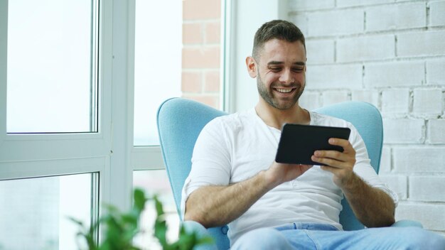Привлекательный улыбающийся мужчина с цифровым планшетом сидит в кресле на балконе в современной квартире на чердаке