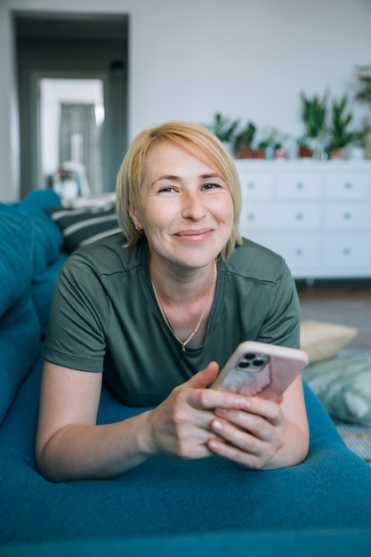 Attractive smiling middle age senior woman using smart phone while is lying on the sofa at home