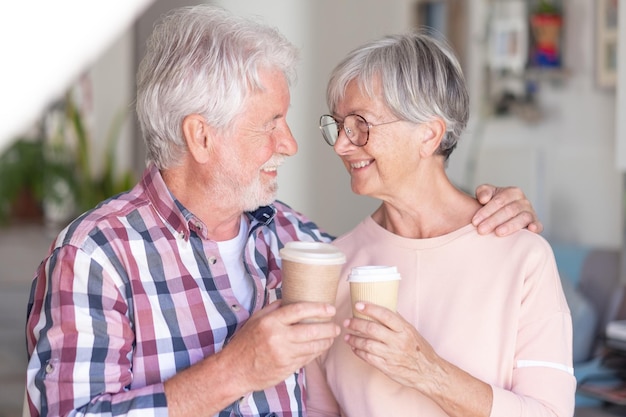 魅力的な笑顔の年配のカップルが2杯のコーヒーを持って目を抱き締めて一緒に快適な2人の白人の先輩のための強い関係