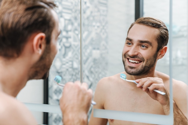 Attraente uomo a torso nudo sorridente che si lava i denti mentre si trova allo specchio del bagno