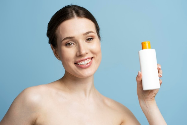 Attractive smiling woman holding shampoo bottle looking at camera isolated on blue background